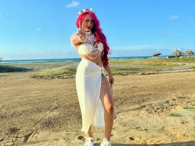 VictoriiaGrey, com cabelo ruivo, cropped branco e saia branca envelope, em pé em uma praia, usando tênis brancos, uma coroa de flores e um colar, apontando para frente com um sorriso confiante..