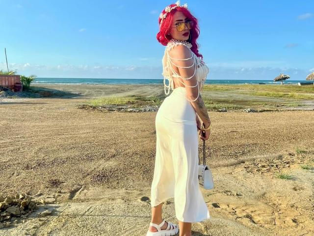 VictoriiaGrey with red hair, white dress, and straps, showing her ass, holding a white bag, standing on a sandy beach, wearing white sandals, and a flower headpiece.