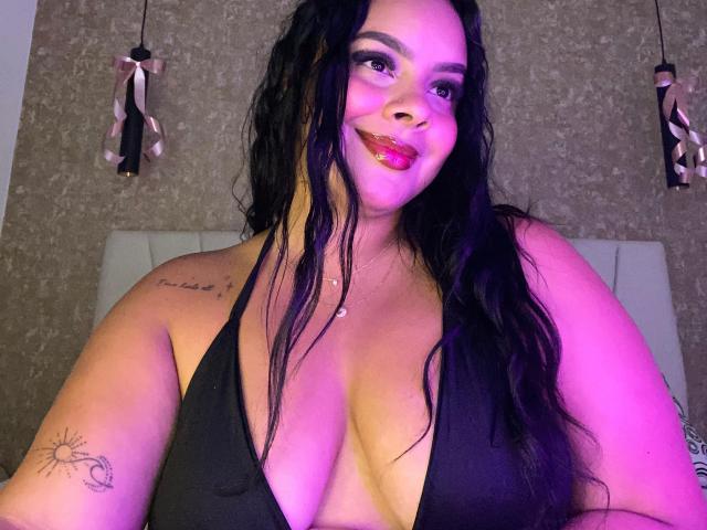 AmarantaCarey with long black hair, pink lipstick, and a black halter top showing her big tits. She has a tattoo on her left arm and a necklace.