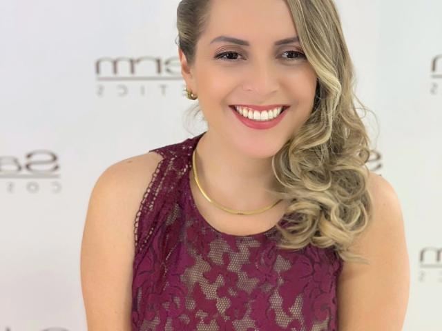 Jammyx69 with blonde, wavy hair, smiling, wearing a lace, sleeveless, burgundy dress, gold necklace, and earrings, showing white teeth and pink lipstick.