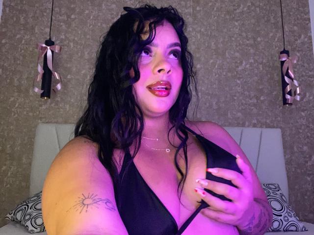 AmarantaCarey with curly black hair, wearing a black halter top, holding her tits, pink lipstick, tattoo on her left arm, and pink ribbons hanging in the background.