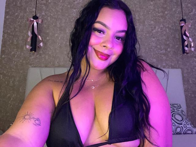 AmarantaCarey with long black hair, purple highlights, smiling, wearing a black bra, showing big tits, sun tattoo on right arm, pink lipstick, purple light, and a necklace.