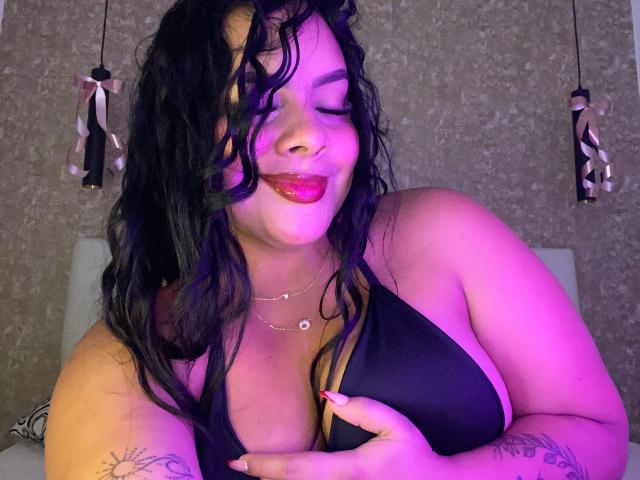 AmarantaCarey with wavy black hair, purple and pink lights, smiling with red lipstick, holding black bra, showing large tits, tattoos on arms, gold necklace, pink ribbons in background.