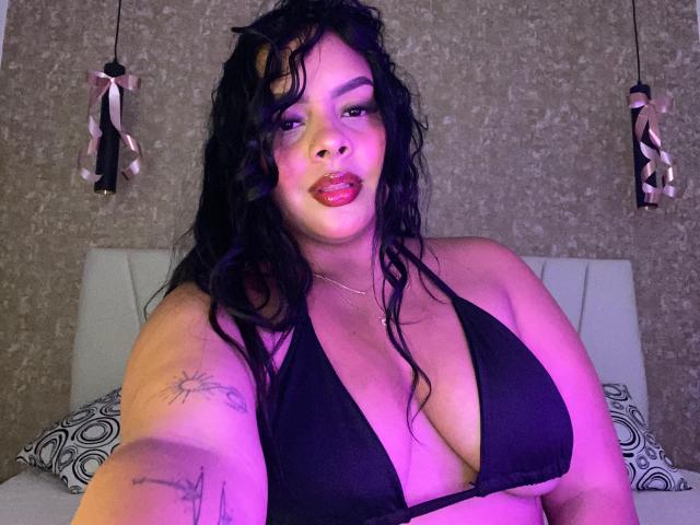 AmarantaCarey with wet black hair, large tits in a black bikini, purple lighting, red lips, tattoos on right arm, and pink ribbons in the background.