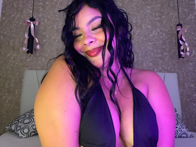 AmarantaCarey with wavy black hair, green eyes, and red lipstick, smiling with eyes closed, wearing a black halter top that reveals large, sexy boobs, in purple lighting.