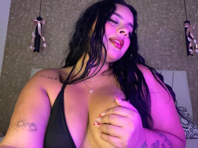 AmarantaCarey with long black hair, purple-lit face, red lipstick, black tank top, hand on big tits, tattoos on arm and chest, ring on finger, confident smile, wavy hair.