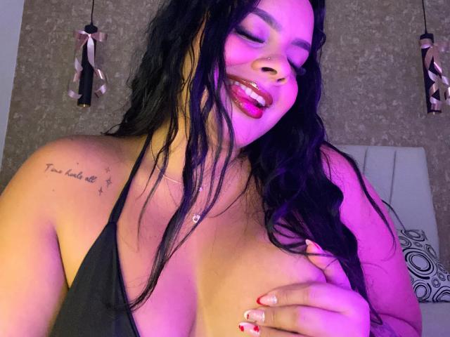 AmarantaCarey with long black hair, pink lips, and a tattoo on her left shoulder, covering her tits with one hand, under purple and pink lighting, showing a playful expression.