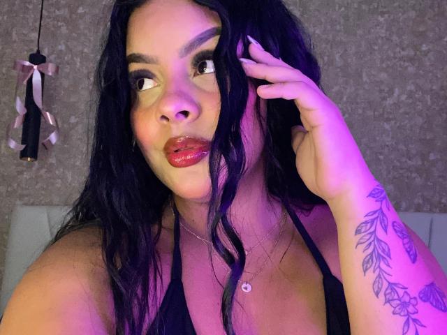 AmarantaCarey with black wavy hair, red glossy lips, black halter top, tattooed arm, purple-pink lighting, hand in hair, looking sideways, necklace with small circular pendant.