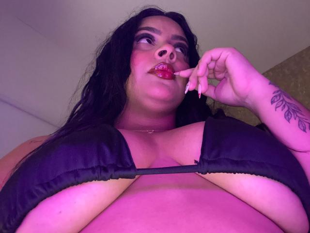 AmarantaCarey with dark hair, pink lipstick, black bra, showing large tits, purple light, tattoo on right arm, biting red lipstick, looking up, low angle shot.