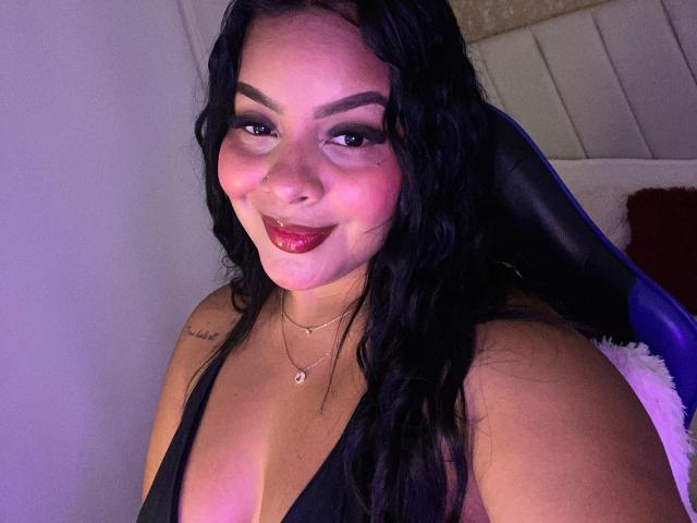 AmarantaCarey with wavy black hair, dark makeup, and red lipstick, smiling at the camera. She's wearing a black top, showing her tits, with a small necklace.