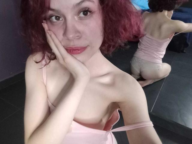 Sophie Blake mit roten Locken, heller Haut und rosa Dessous, Hand am Wangen, blickt in einen Spiegel, der ihren nackten Hintern, ihre kleinen Brüste und die rosa Unterwäsche zeigt, in einem schwach beleuchteten Raum..