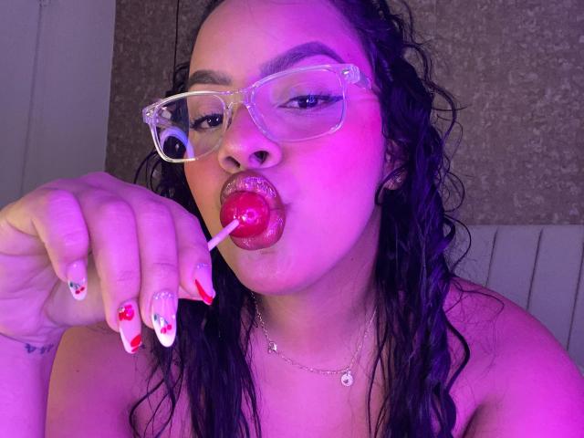 AmarantaCarey with wet black hair, wearing clear glasses, sucking a red lollipop, has red nail polish with white patterns, and a necklace, under purple lighting.