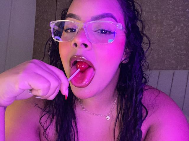 AmarantaCarey with wet black hair, wearing transparent glasses, sucking a red lollipop, tongue out, red nails, necklace, and purple-pink lighting. Boobs visible.