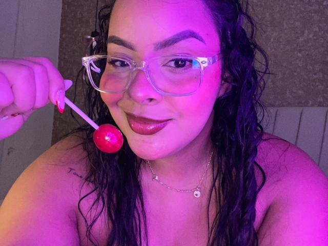 AmarantaCarey with wet black hair, wearing clear glasses, holding a red lollipop near her glossy pink lips, and a necklace, has a tattoo on her right shoulder.