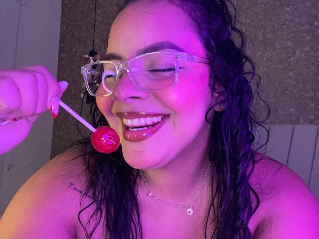 AmarantaCarey with wet black hair, clear glasses, glossy pink lips, smiling, holding a red lollipop near her face, pink and purple lighting, visible tattoo on right shoulder.