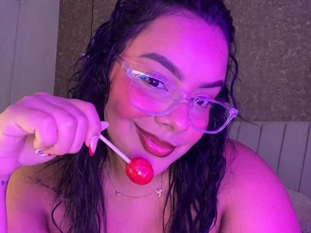 AmarantaCarey with wet black hair, clear glasses, and red lollipop, smiling with glossy pink lips, holding lollipop near mouth, wearing a necklace, in purple-lit photo.