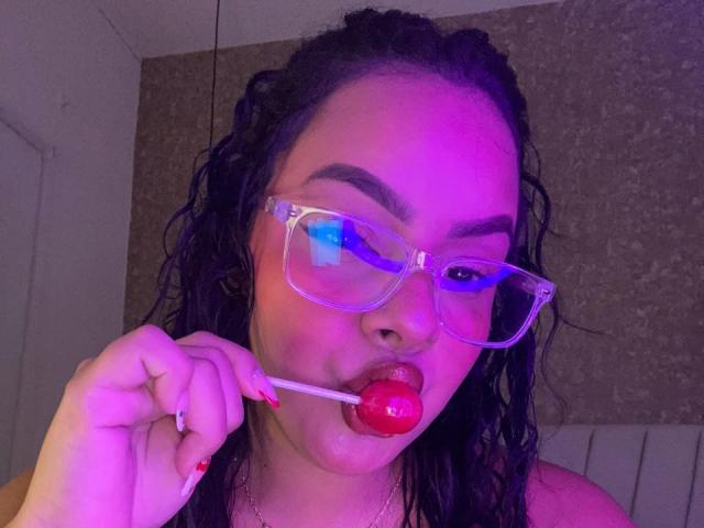 AmarantaCarey with wet black hair, purple neon light, and clear glasses, sucking a red lollipop, showing pink lipstick and red nails, looking cute and sexy.