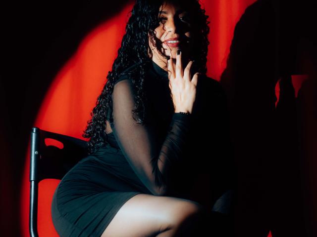 AlissaaMilf with curly black hair, wearing a black sheer dress, sits on a chair, touching her lips with her finger, red light casting shadows on her face.