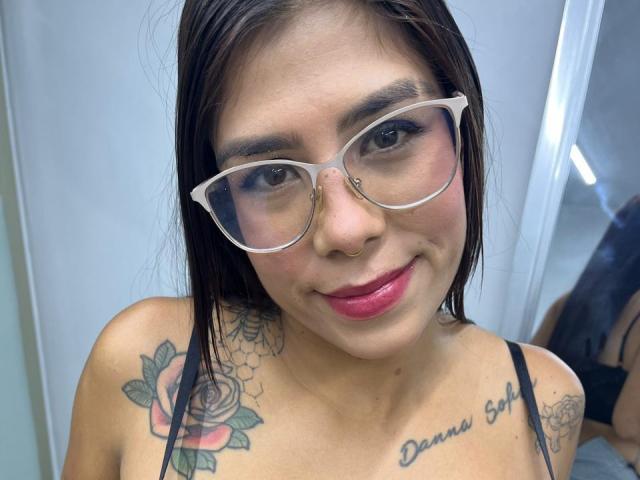 VioletaGates with glasses, pink lipstick, and brown hair, smiling at the camera. She has tattoos of roses and text on her chest, and is wearing a black strap top.
