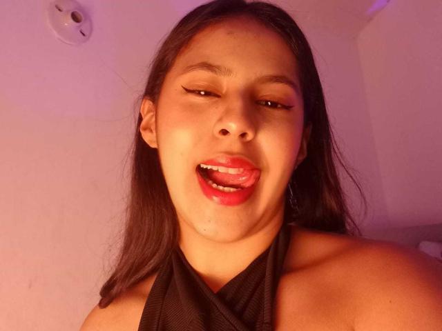 SoffiRousee with dark hair, brown eyes, and red lipstick, sticking out her tongue, wearing a black halter top, and having visible drool on her chin.