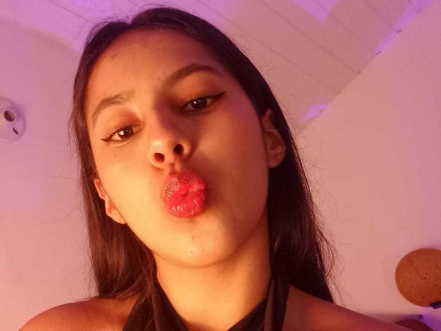 SoffiRousee with pouty, pink, glossy lips, long dark hair, brown eyes, and subtle makeup, wearing a black halter, in a warm, purple-lit room.