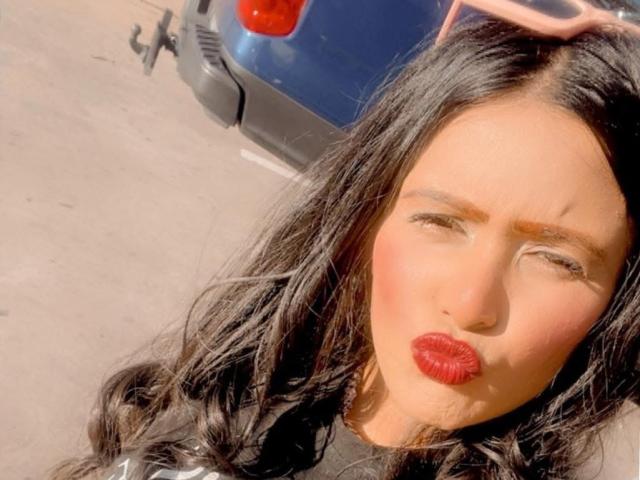 IndiaPrincess has sunburned face, black wavy hair, red lipstick, and a pouty expression with a pink headband. She's in front of a blue car.