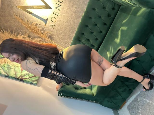 AmaraRivero with long brown hair, black fishnet arm sleeves, tight black leather skirt, and black high heels, leaning on a green tufted couch, showing her big ass and tattoo.