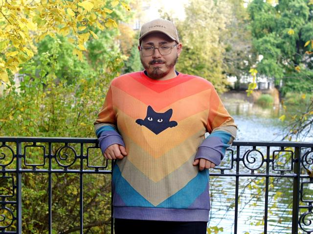 FabianNoLimits, chubby dude with glasses, beige cap, rainbow sweater with cat face, standing by black fence, hands on bars, autumn foliage, green and yellow leaves, water in background.