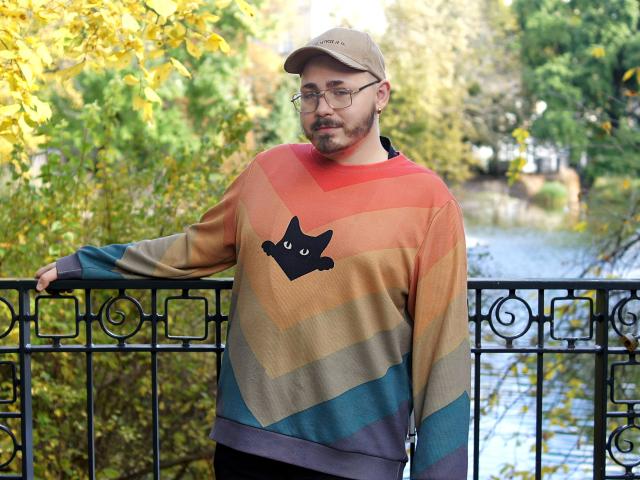 FabianNoLimits, chubby dude with glasses and beard, wearing a rainbow-striped sweater with a black cat face, beige cap, standing by a black iron railing, looking serious.