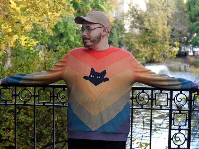 FabianNoLimits with glasses, beard, and cap, wearing a rainbow sweater with a black cat face, arms out, leaning on a black iron railing, standing by a river.