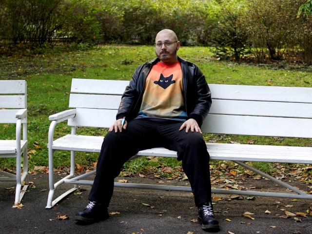 FabianNoLimits, bald, with glasses, sits on a white bench in a park, wearing a black leather jacket, orange and black cat shirt, black pants, and black sneakers.