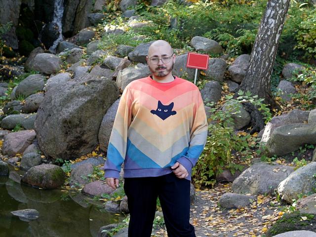 FabianNoLimits, bald with glasses, beard, wearing a colorful sweater with a black cat face, black pants, standing by a rocky pond, forest background, autumn leaves.