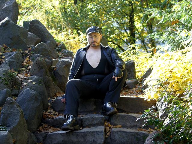 FabianNoLimits, a bearded dude with a black leather jacket, cap, and fingerless gloves, sits on stone steps, wearing black pants and boots, surrounded by rocks and autumn leaves.