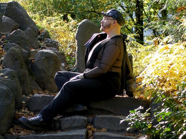 FabianNoLimits, fat dude with a black cap, brown puffy jacket, and black pants, sitting on rocks in a sunny forest, looking up, backpack on, boots on.