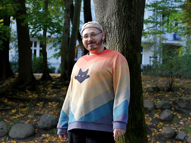 FabianNoLimits, a chubby bearded dude with glasses, wearing a rainbow sweater with a cat face, and a beige headscarf, standing against a tree in a forest.