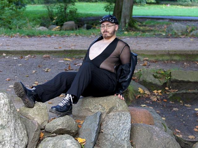 FabianNoLimits, chubby, wearing sheer black outfit, black boots, and cap, sitting on rocks, showing chest, looking serious, autumn park, white laces, black leather jacket.