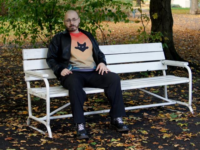 FabianNoLimits, bald with glasses, black jacket, beige shirt with cat design, black pants, black shoes, sitting on white bench, autumn leaves on ground, relaxed, right hand on lap.