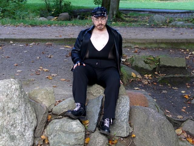 FabianNoLimits, a chubby dude with a beard, black leather jacket, see-through black top, black pants, black boots, and a black cap, sitting on rocks in a park.
