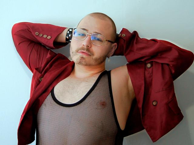 FabianNoLimits, bald with a beard, wearing glasses, red jacket, black mesh top, arms raised, showing chest and nipples, black wristband, light skin, confident pose.