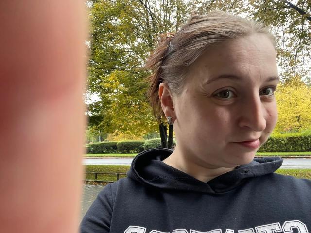 Carmen-hot has pale skin, brown eyes, and a slight smile. She's wearing a black hoodie, with brown hair in a messy bun, and small earrings. A blurry pink object is in the foreground.