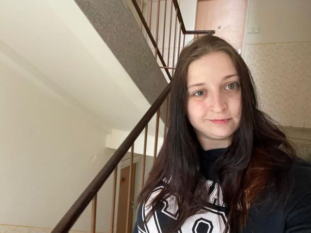 Carmen-hot has pale skin, long brown hair, and green eyes. She's wearing a black and white sports jersey, standing near a stair railing with a subtle smile.