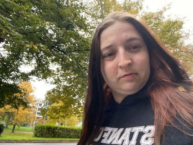 Carmen-hot with long brown hair, pale skin, and a neutral expression, wearing a black hoodie, looking slightly down at the camera. Her tits are not visible.