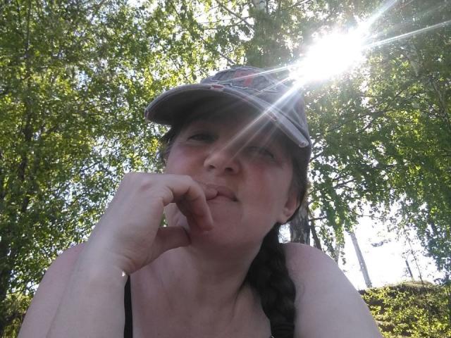 IllusionGold, avec une expression pensive, la lumière du soleil filtrant à travers une casquette, un doigt touchant ses lèvres, un débardeur noir et des cheveux tressés, entourée d'arbres verts..