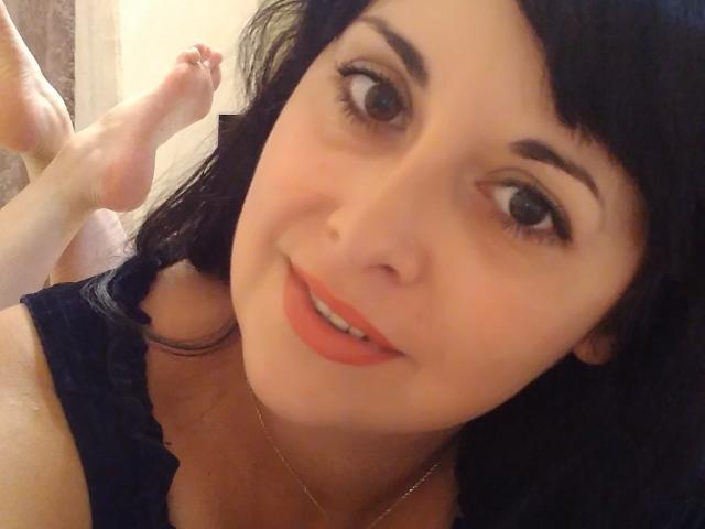 Close-up selfie of SelieAlMio with dark, wavy hair and bangs. She has fair skin, brown eyes with long lashes, and a subtle smile with pink lipstick. She's wearing a dark blue, lace-trimmed top. Her feet are raised in the background. The image has a soft, warm light, highlighting her gentle expression. The focus is on her face, with her eyes looking slightly to the right. She has a thin gold necklace.