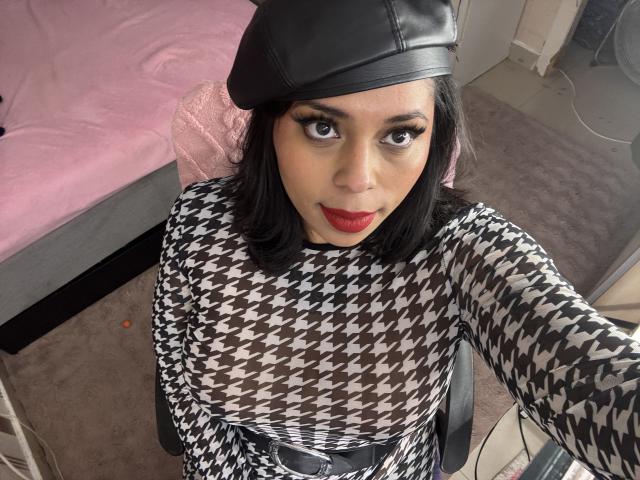 Chili, with black hat and houndstooth dress, gazes up, red lips, black chair, bed in pink background.