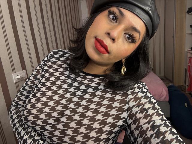 Chili has black hair, red lipstick, black-and-white houndstooth shirt, black leather cap, gold earrings, looking at camera, striped wall.