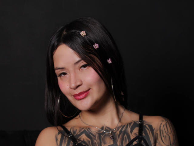 Parisnoa with black hair, pink flower hairpins, hoop earrings, red lipstick, tattooed chest, smiling, dark background, medium skin, cute, sexy, looking at camera.