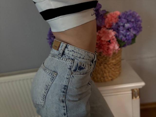 Amabel's ass in light blue denim jeans, slightly pulled down, showing a sliver of her white and black top. Woven vase with pink and purple flowers on a white table.
