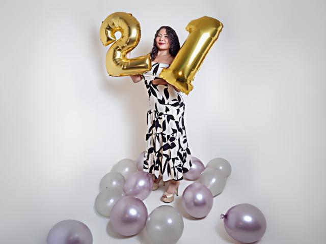 JadeFoxx hält glänzende, goldene "21"-Ballons, trägt ein weißes und schwarzes Kleid mit Muster und steht auf einer Ansammlung von weißen und rosafarbenen Ballons..