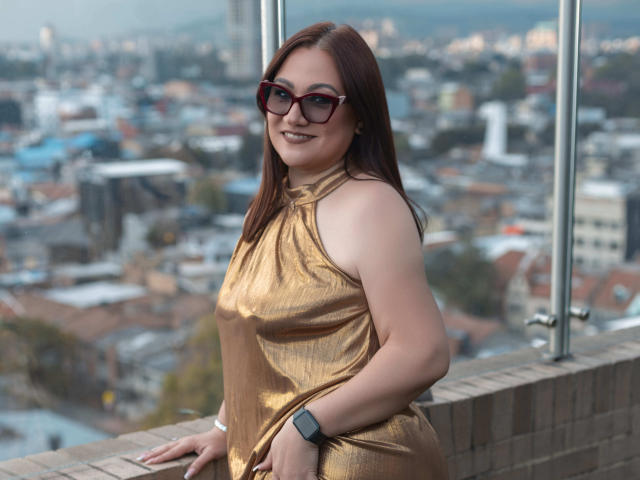 LadyNylonOh with red hair, glasses, gold halter dress, and wristwatch, standing by a glass balcony, showing her ass slightly, cityscape blurred in the background.